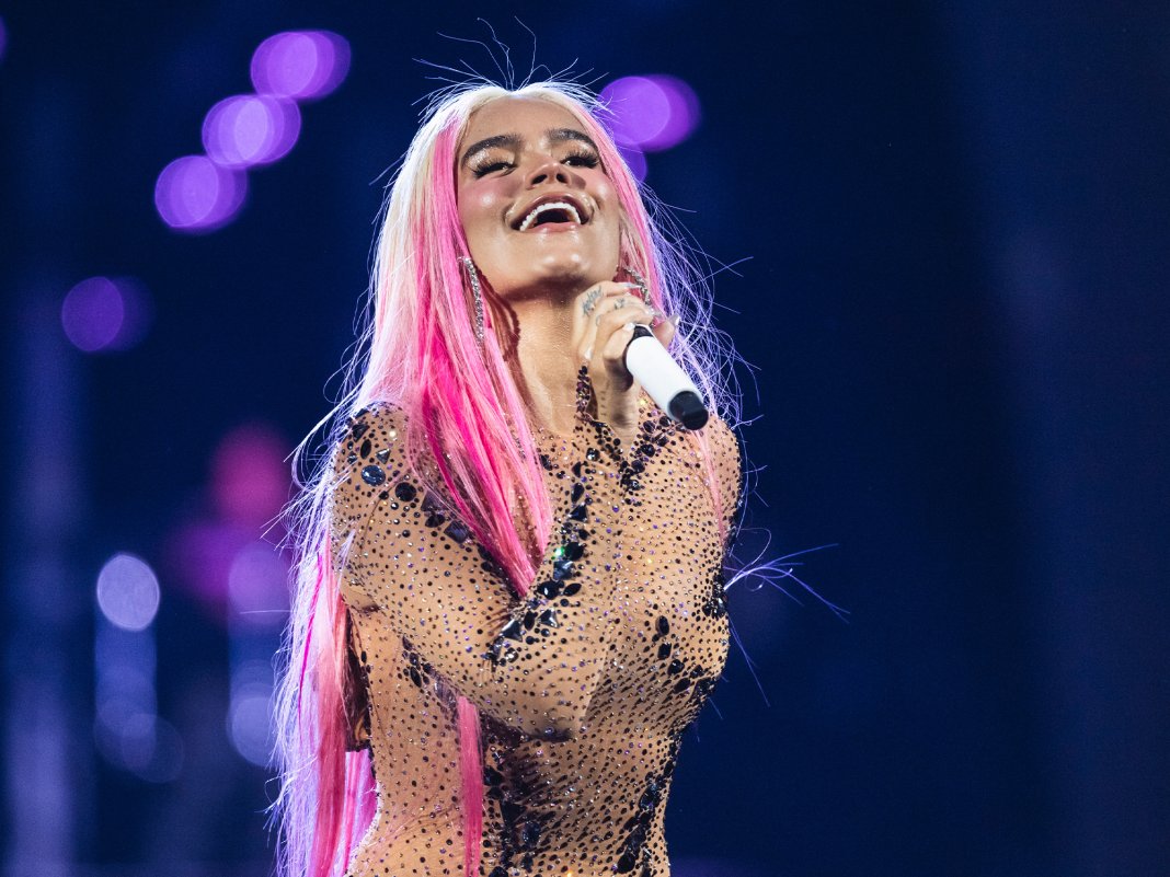 Karol G with a mic in hand, pictured smiling as she is singing.