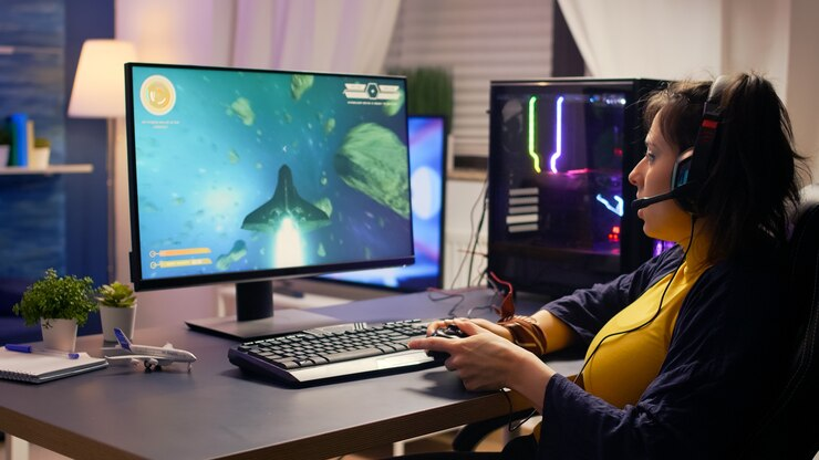 Person wearing a headset playing a space-themed video game on a computer, with a keyboard, mouse, and gaming controller on the desk.