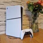 Test de la PS5 Slim : plus fine, mais toujours puissante The PS5 Slim and DualSense controller on a wooden table in front of a white brick background and next to a vase of flowers